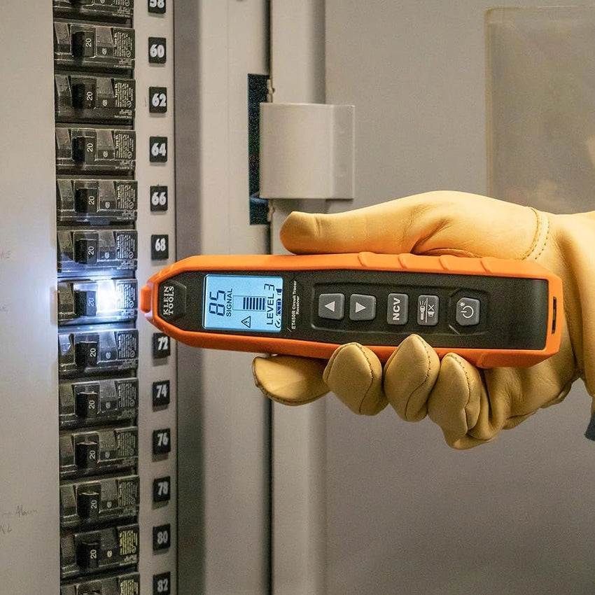Hand wearing a yellow glove holding an orange digital device in front of an electrical panel.
